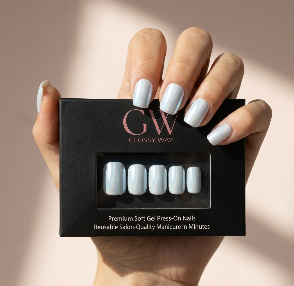 A hand holding a black box of glossy press-on nails, with the nails themselves shown through the front window. The nails have a pale icy blue-gray glossy finish, and the background is a soft warm beige with natural shadows.