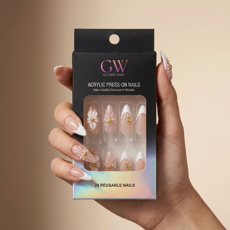A hand holds a black box of GW Closely Waw acrylic press-on nails. The nails are shown through the clear window on the packaging, featuring a nude pink base with white French tips, delicate gold glitter details, and pearl accents.