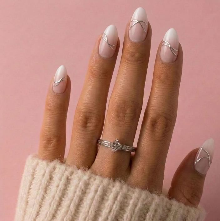 A close-up of a hand with long almond-shaped nails painted in a glossy white French tip style with thin silver curved line details. A silver ring is worn on one finger, and the hand is wearing a cream-colored knit sweater against a solid pink background.