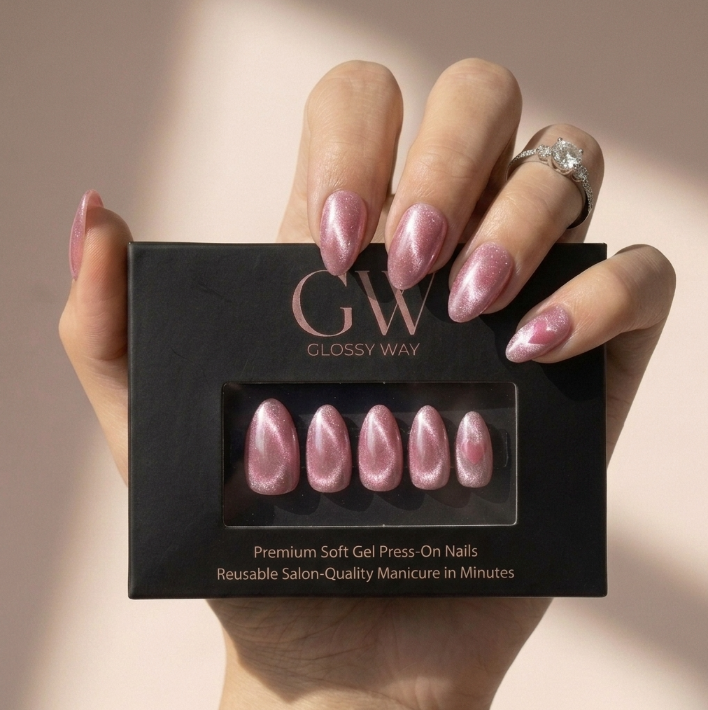 A hand holding a black Glossy Way press-on nail box, showing six pink metallic press-on nails inside. The nails have a shimmering cat-eye finish, and the hand is wearing a silver ring against a soft beige background.