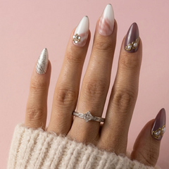 A close-up of a hand with long almond-shaped nails decorated in elegant nail art, wearing a silver ring with clear stones. The nails feature white and pink marbled designs, a metallic silver leaf pattern, and gold leaf accents, set against a soft pink background.