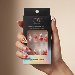 A hand holding a black glossy press-on nails box labeled 'GW GLOSSY WAY' and 'ACRYLIC PRESS-ON NAILS' with a window showing sample press-on nails. The nails are white with pink gradient tips and floral designs, set against a warm beige background.
