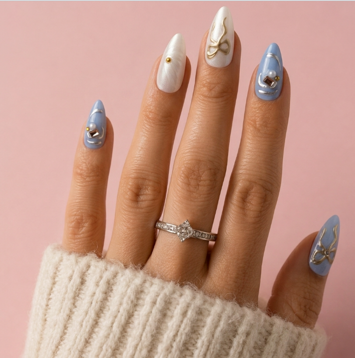 A close-up of a hand with long almond-shaped nails styled in a pastel manicure. The nails feature white with gold bow designs, light blue with gold leaf designs, and one light blue nail with a small gold heart.