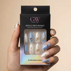 A hand holds a black box of GW Glossy Way acrylic press-on nails against a warm beige background. The nails shown through the window are white and light blue with gold glitter swirls and small rhinestone accents.