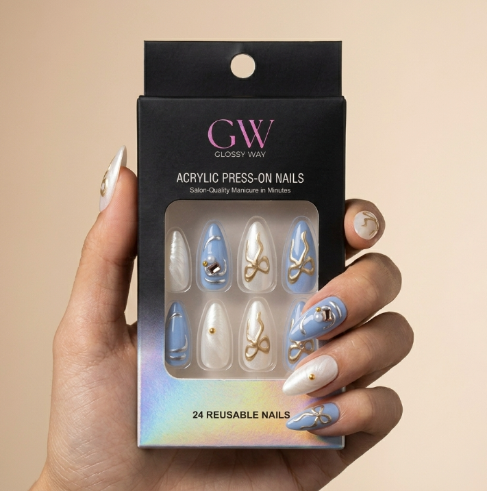 A hand holds a black box of GW Glossy Way acrylic press-on nails against a warm beige background. The nails shown through the window are white and light blue with gold glitter swirls and small rhinestone accents.