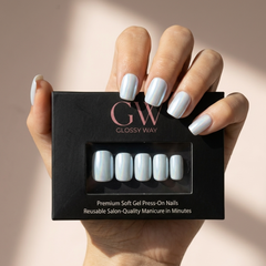 A hand holding a black box of glossy press-on nails, with the nails themselves shown through the front window. The nails have a pale icy blue-gray glossy finish, and the background is a soft warm beige with natural shadows.