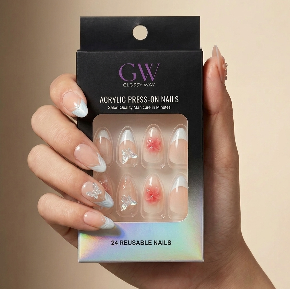 A hand holding a black glossy press-on nail package with a clear window showing 24 reusable acrylic nails. The nails have a sheer nude base with white French tips, clear rhinestone embellishments, and two pink floral designs.