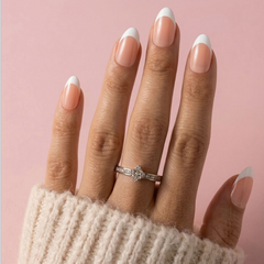 A close-up of a hand with French tip manicure wearing a silver diamond ring, set against a soft pink background.
