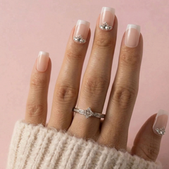 A close-up of a hand wearing a silver ring with a clear central gemstone and smaller side stones, against a soft pink background. The nails are styled with a pale pink and white French manicure and small rhinestone accents.