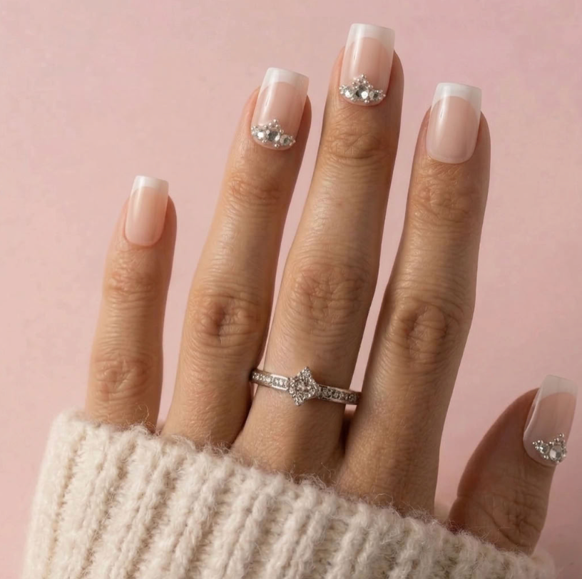 A close-up of a hand wearing a silver ring with a clear central gemstone and smaller side stones, against a soft pink background. The nails are styled with a pale pink and white French manicure and small rhinestone accents.