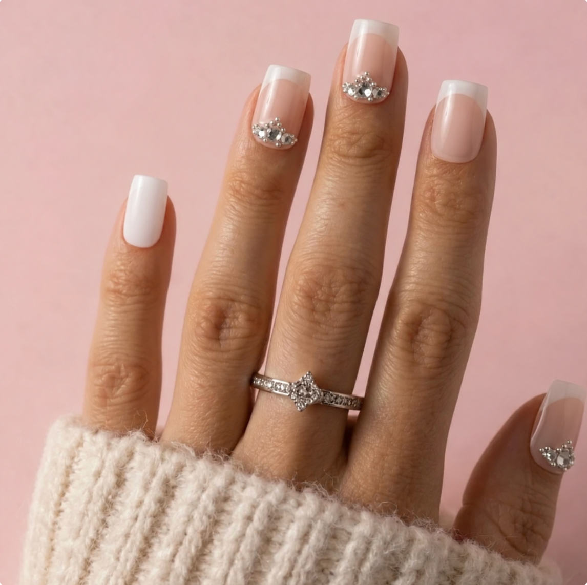 A close-up of a hand with a French manicure, wearing a silver diamond ring on one finger and a cream knit sweater on the wrist, set against a solid pink background.