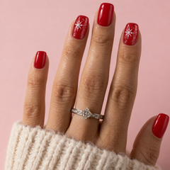 A close-up of a hand wearing a silver ring with a clear center stone, decorated with red nail polish and white star designs, set against a solid pink background.