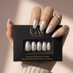 A hand holding a black box of Glossy Way press-on nails. The nails are glossy white with delicate silver glitter swirls and small rhinestone accents.