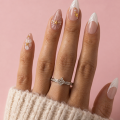 A close-up of a hand with long almond-shaped nails styled in a French tip and bohemian-inspired manicure, wearing a silver diamond ring and a cream knit sweater against a solid pink background.