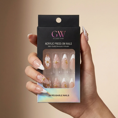 A hand holds a black box of GW Closely Waw acrylic press-on nails. The nails are shown through the clear window on the packaging, featuring a nude pink base with white French tips, delicate gold glitter details, and pearl accents.