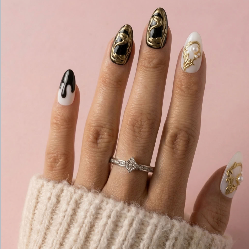 A close-up of a hand showcasing a set of almond-shaped press-on nails with three distinct designs: black and white dripping, metallic gold abstract swirls, and white with gold floral details. A silver ring is worn on the middle finger, and the hand is resting on a cream knit sleeve against a solid pink background.