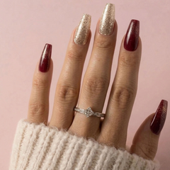 A close-up of a hand with long coffin-shaped nails styled in a holiday palette, featuring two glittery gold nails and two deep burgundy ombre nails. A silver diamond ring is worn on the ring finger, and the hand is wrapped in a cream-colored knit sweater against a soft pink background.