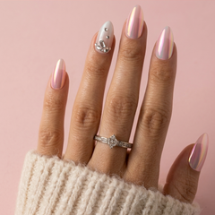 A close-up of a hand with long, glossy, iridescent pink almond-shaped nails, wearing a silver ring with a clear floral center stone. The hand is wrapped in a cream-colored knit sweater against a solid light pink background.