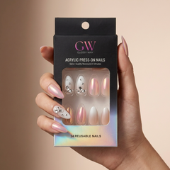 A hand holding a black box of GW Glossy Way press-on nails. The nails are shown through the clear window on the packaging, featuring a mix of metallic rose gold, pearlescent white, and silver rhinestone designs.