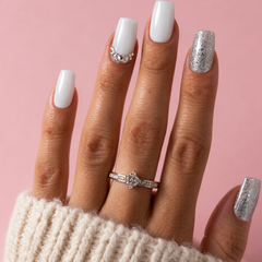 A close-up of a hand with white and silver manicured nails, wearing a silver ring with a clear marquise-cut stone, set against a solid pink background.