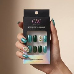 A hand holds a black box of GW Glossy Way acrylic press-on nails. The nails are arranged in a clear window and feature a teal glitter design with green, dark green, and white glitter/nail art designs.
