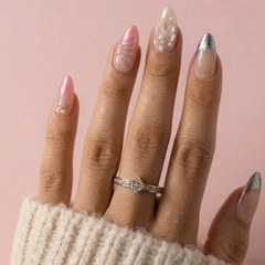 A close-up of a hand with long almond-shaped nails decorated in pastel pink, white, and metallic silver designs, wearing a silver ring with clear stones, set against a soft pink background.