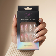 A hand wearing a beige knit sweater holds a black box of GW Glossy Way press-on acrylic nails. The nails are shown through a clear window on the packaging, featuring pink, nude, and white designs with pearl and glitter accents.