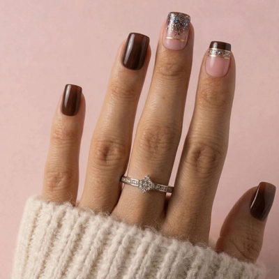 A close-up of a hand with manicured nails in shades of brown, pink, and glitter, wearing a silver ring with clear stones on the ring finger, set against a soft pink background.