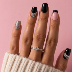 A close-up of a hand with manicured nails in black, pale pink, and glittery silver, wearing a silver ring with a clear pavé center, set against a solid pink background.