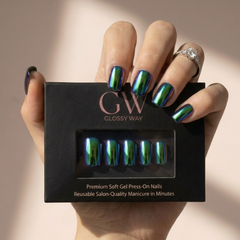 A hand holding a black box of Glossy Way press-on nails, with the nails displayed in a clear window on the box. The nails have an iridescent, holographic finish shifting between green, blue, and purple.
