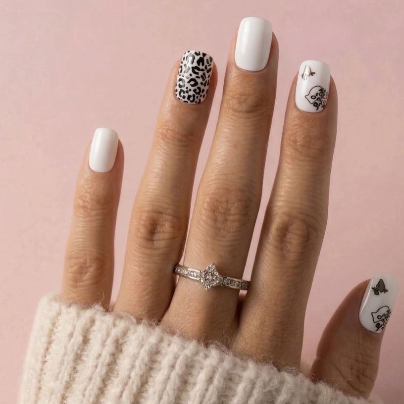 A close-up of a hand wearing a silver ring with a clear center stone, featuring white, black leopard print, and black butterfly nail art on a soft pink background.