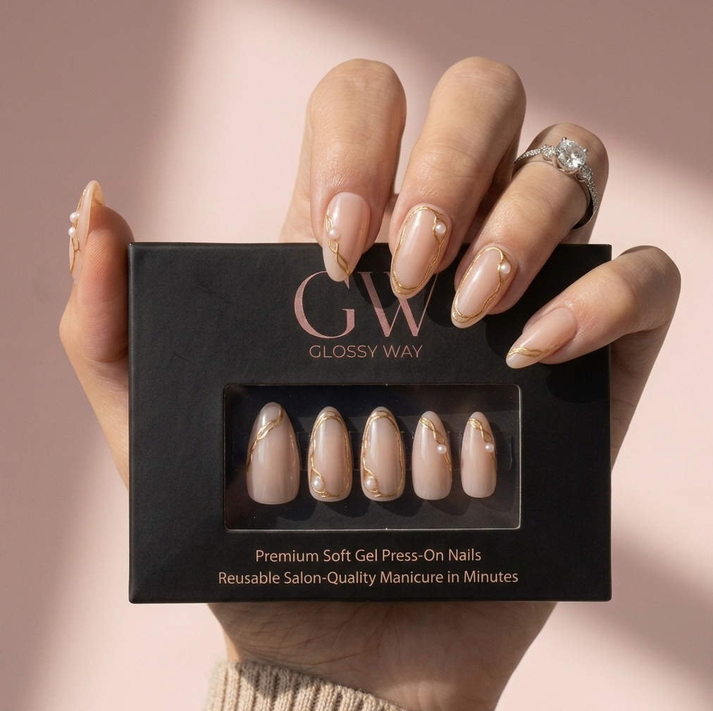 A hand holding a black glossy press-on nails box with five nude beige nails featuring gold glitter and pearl details visible through the clear window. The hand has matching nude manicured nails and a silver ring on one finger, set against a soft pink background.