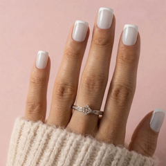 A close-up of a hand with white French-tip nails wearing a silver diamond ring with a starburst center, set against a soft pink background.