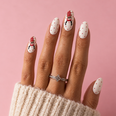 A close-up of a hand with manicured nails painted white with small red polka dots and two snowman illustrations, wearing a silver ring with clear stones and a cream knit sleeve against a pink background.