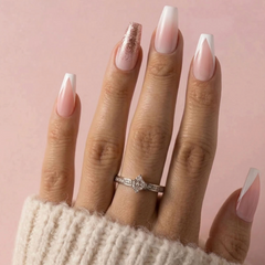 A close-up of a hand with long almond-shaped nails featuring a French tip design in white and soft pink, with one glittery pink nail. A silver ring is worn on the ring finger against a solid pink background.