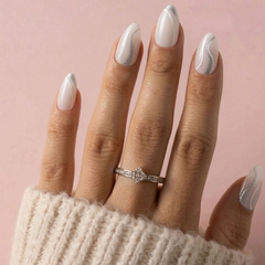 A close-up of a hand with long white stiletto nails decorated with silver wavy lines, wearing a silver diamond engagement ring. The hand is resting on a soft pink background with a cream knit sleeve visible at the bottom.