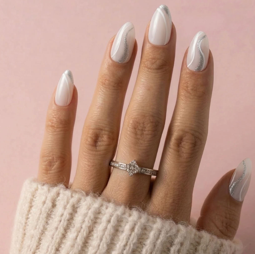 A close-up of a hand with long white stiletto nails decorated with silver wavy lines, wearing a silver diamond engagement ring. The hand is resting on a soft pink background with a cream knit sleeve visible at the bottom.