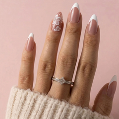 A close-up of a hand with almond-shaped French tip nails, featuring a nude pink base with white tips and one white floral nail art design. A silver ring with a central clear gemstone is worn on the ring finger against a soft pink background.