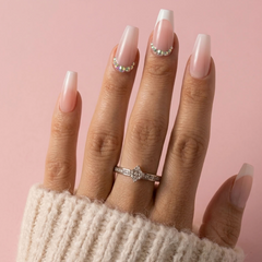 A close-up of a hand with long, square-shaped nails styled in a soft pink gradient with white tips and small rhinestone accents. The hand is wearing a silver ring with a clear central stone, and a cream-colored knit sleeve is visible at the wrist, set against a solid pink background.