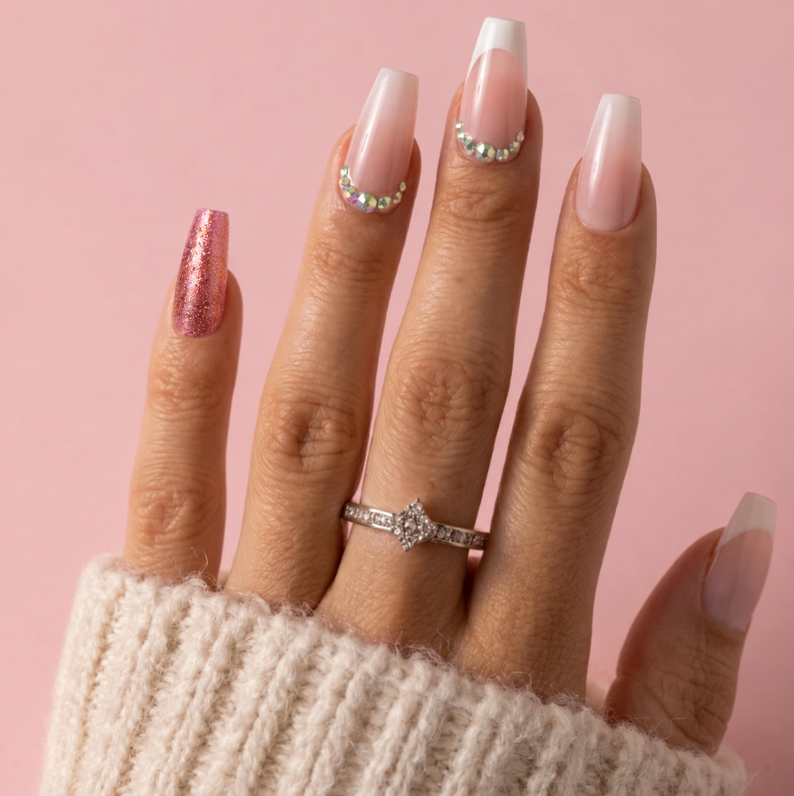 A close-up of a hand with long, glossy almond-shaped nails styled in a French tip and glitter pattern, wearing a silver diamond ring and a cream knit sweater against a solid pink background.