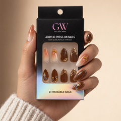 A hand wearing glossy press-on nails holds a black box of GW Glossy Way acrylic press-on nails. The nails feature a gradient peach-to-white design with gold glitter and brown glitter designs.