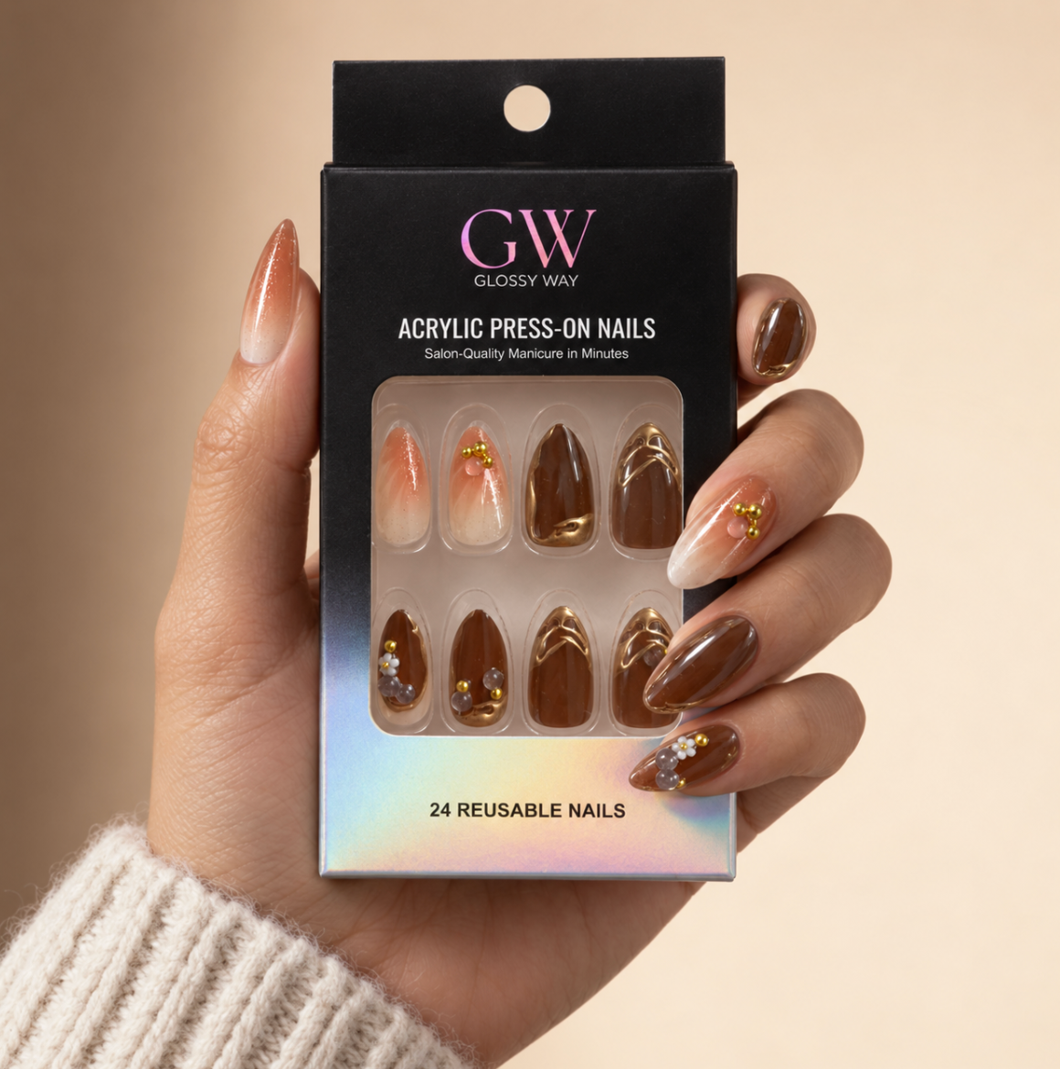A hand wearing glossy press-on nails holds a black box of GW Glossy Way acrylic press-on nails. The nails feature a gradient peach-to-white design with gold glitter and brown glitter designs.