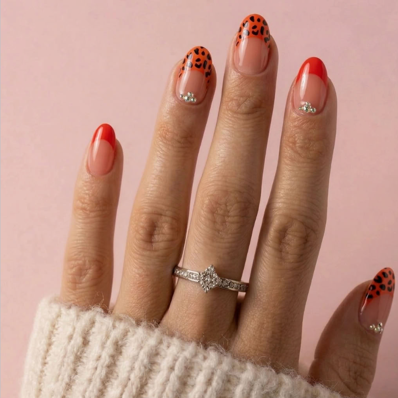 A close-up of a hand with manicured nails featuring a mix of solid red, sheer nude, and orange leopard print designs, wearing a silver ring on the middle finger against a soft pink background.