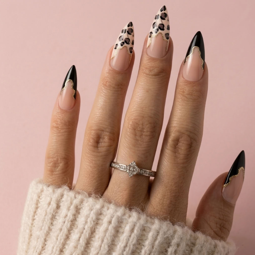 A close-up of a hand with long stiletto-style nails featuring a mix of solid black, nude pink, leopard print, and gold foil accents, wearing a silver ring against a solid pink background.