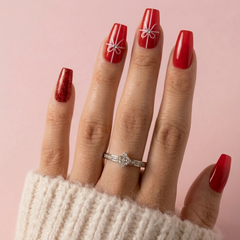 A close-up of a hand with long red manicured nails, decorated with white bow nail art and a glittery red accent nail, wearing a silver diamond ring against a soft pink background.